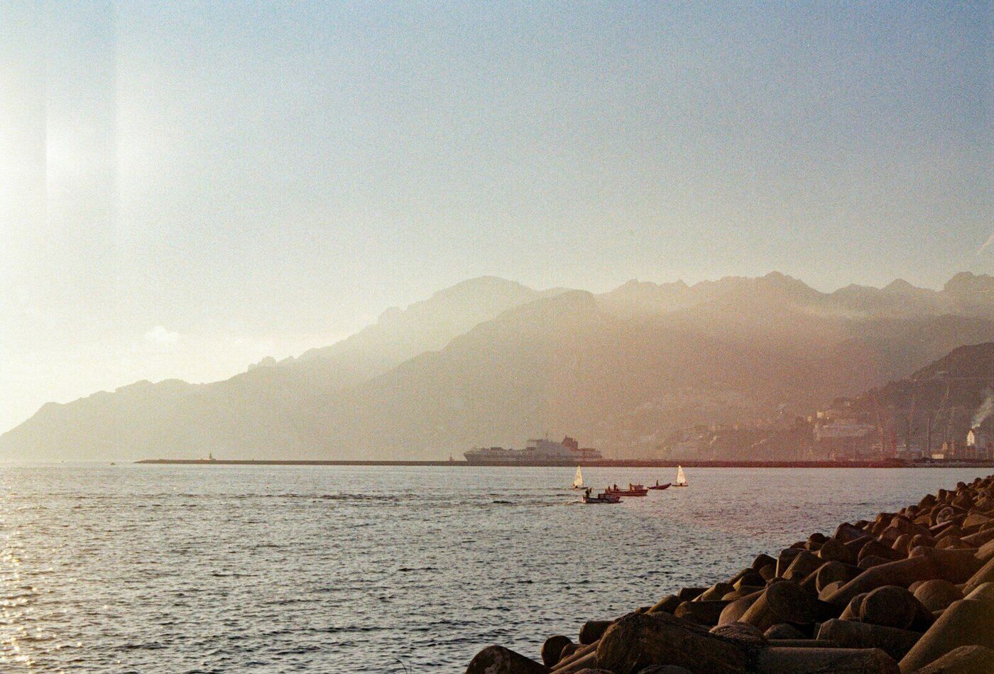 Small sailboats crossing a harbor at golden hour, hazy mountains rising behind the far shore